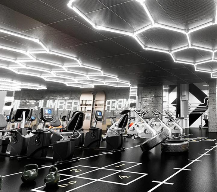 Modern commercial gym floor with hexagonal LED light fixtures on dark ceiling above black rubber floor and cardio equipment