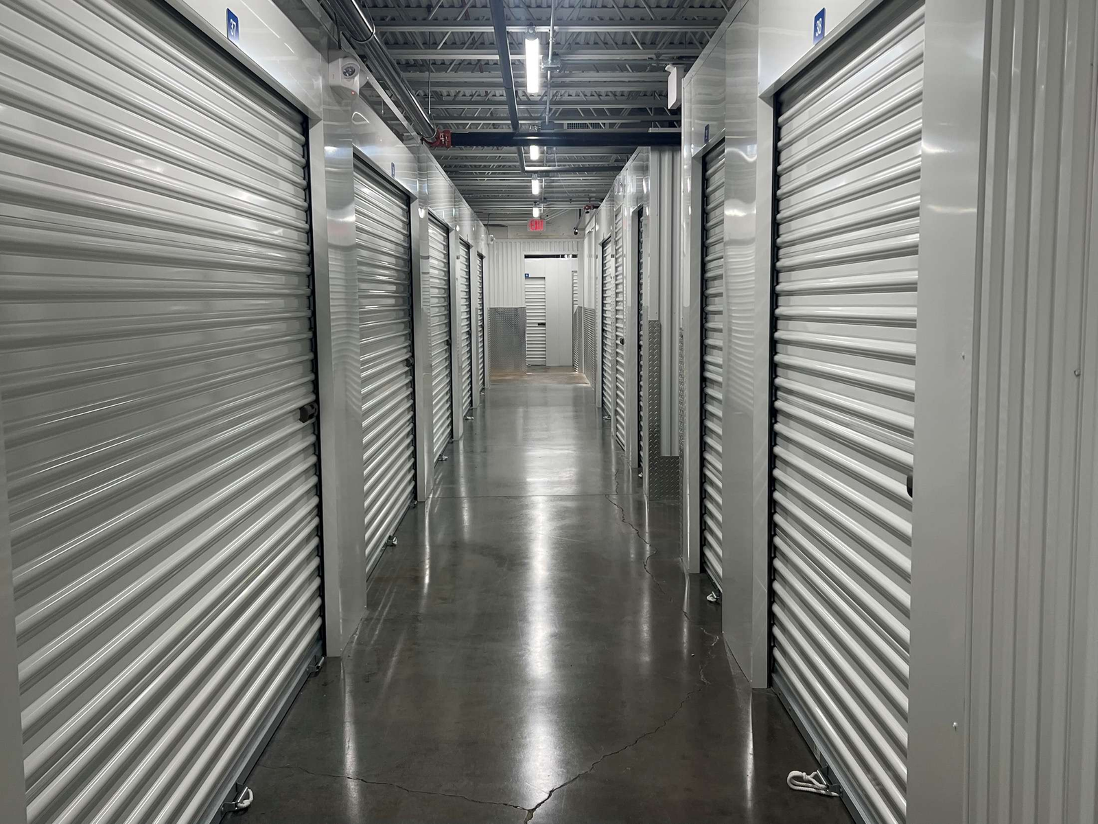 Interior self-storage corridor with white corrugated roll-up doors on both sides, exposed metal ceiling trusses with LED strip lighting fixtures, an EXIT sign visible in the distance, security camera mounted on ceiling, polished gray concrete floor reflecting the overhead LED light