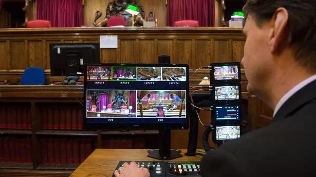 Behind the scenes view of a UK courtroom broadcast control room showing an operator in a dark suit seated at a multi-monitor console with INPUT1, INPUT2, INPUT3, INPUT4 feeds and PGM/PVW preview, with red-curtained courtroom visible in the background
