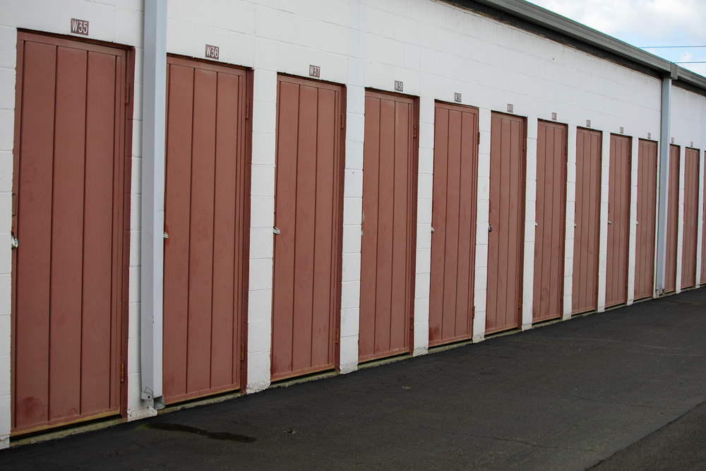 Exterior drive-up self-storage units with red-brown corrugated metal doors numbered W35 through W40 visible on white painted cinder block walls, dark asphalt drive aisle in the foreground, overcast sky, with vertical downspouts between unit groupings