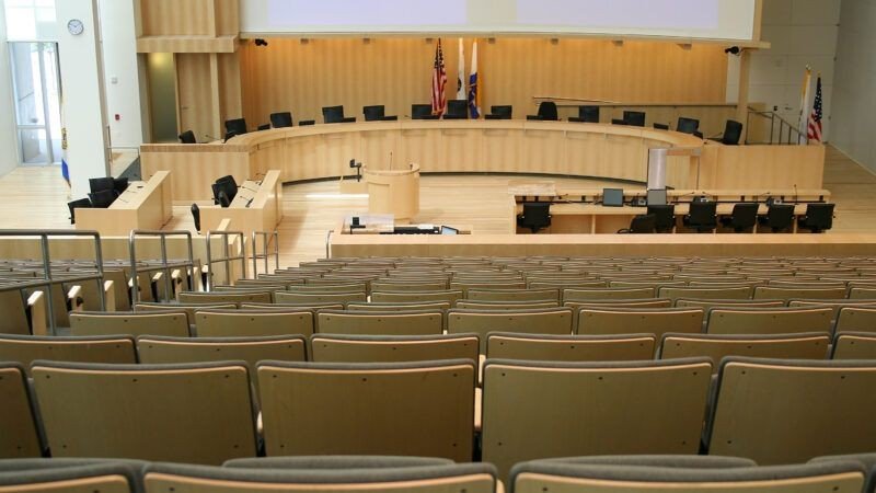 Modern American city council chamber auditorium with a curved wooden dais, multiple black office chairs for council members, US flag and state flag behind the dais, and tiered rows of public seating facing the front where lighting decisions are made