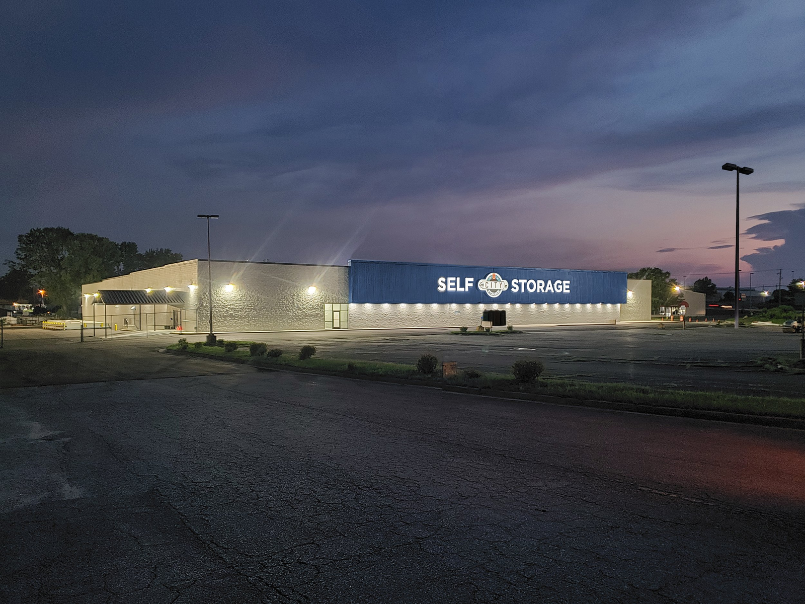 Dusk view of a modern self-storage facility with a blue and white SELF STORAGE sign on the main office building, wall-wash LED lighting on the textured stone facade illuminating the company signage, dark parking lot in the foreground, tall pole lights visible at the right edge of the frame, purple-pink sunset sky